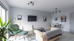 Un salon moderne avec un fauteuil vert, un canapé beige, une table à manger avec des chaises, un téléviseur fixé au mur et une grande fenêtre.