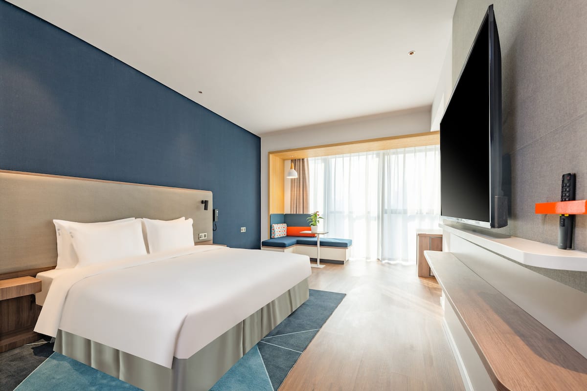 A modern hotel room with a large bed, a flat-screen TV mounted on the wall, a seating area with a table and chairs, and a large window with curtains.