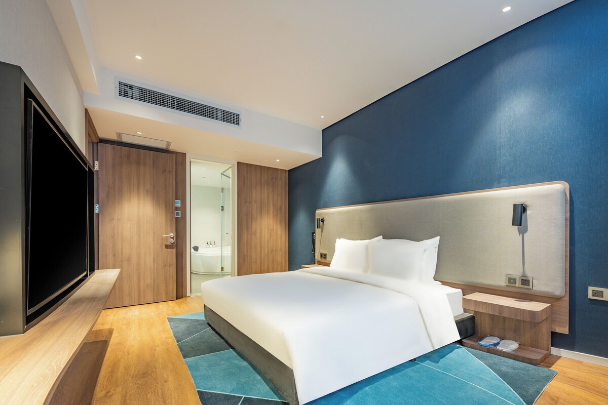 A modern hotel room with a large bed, a flat-screen TV, wooden flooring, and a blue accent wall.