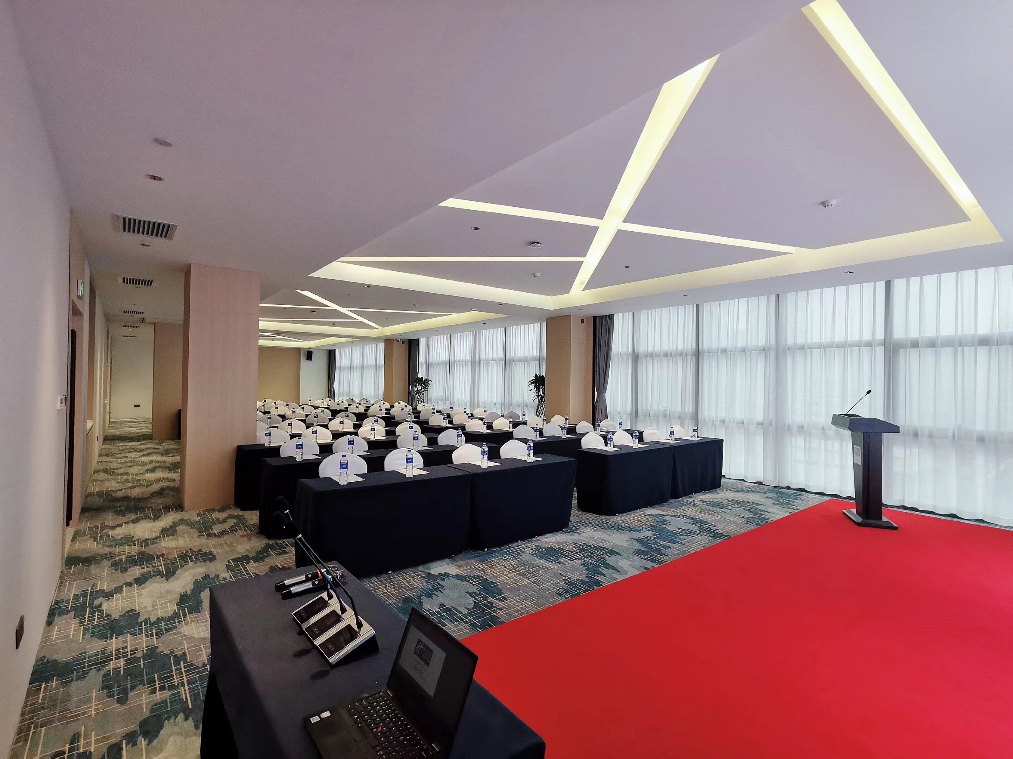 Un salón de conferencias con filas de mesas y sillas, un atril y suelo alfombrado.
