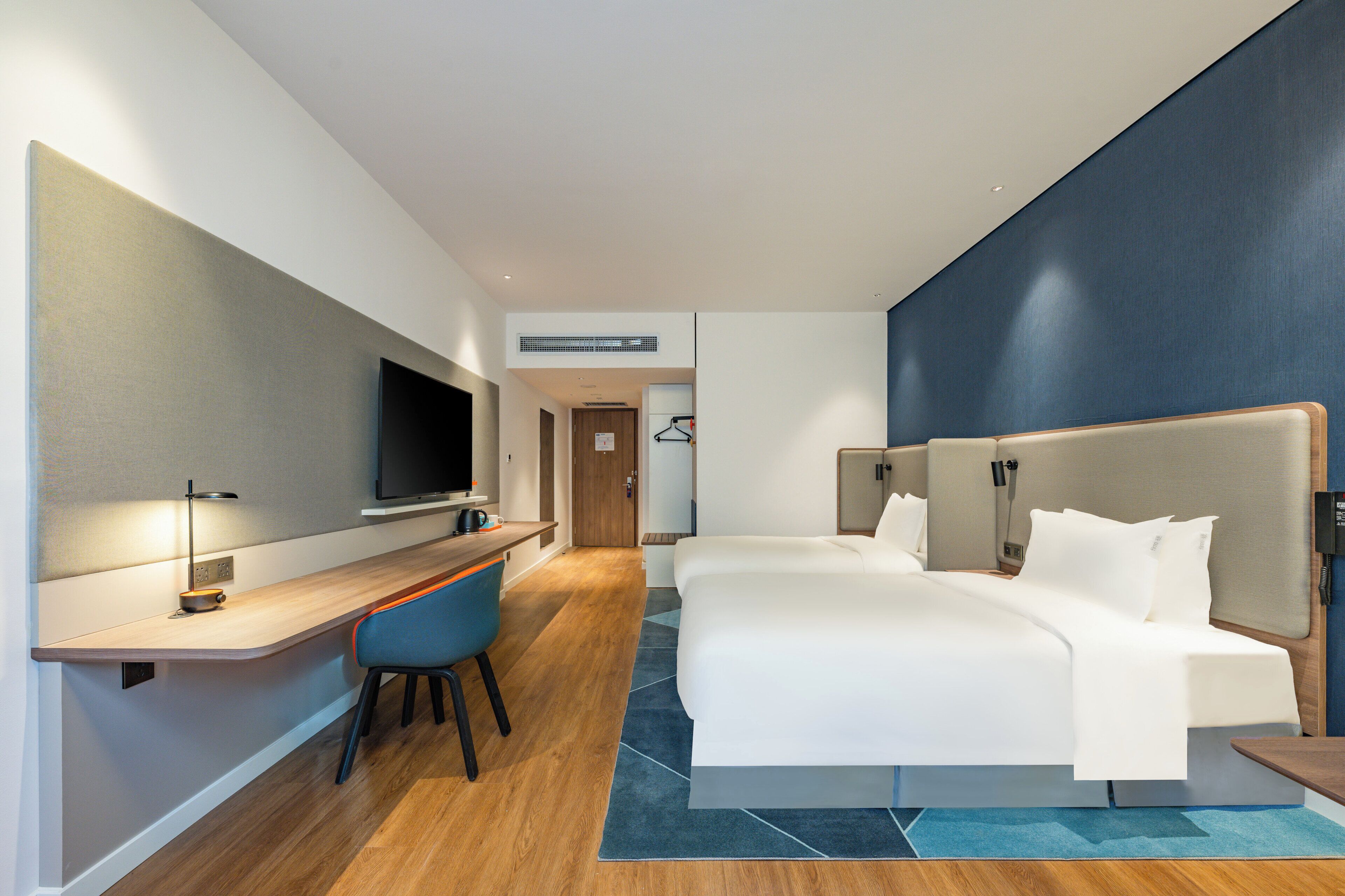 Quarto de hotel moderno com escrivaninha, duas camas, televisão e parede azul decorativa.