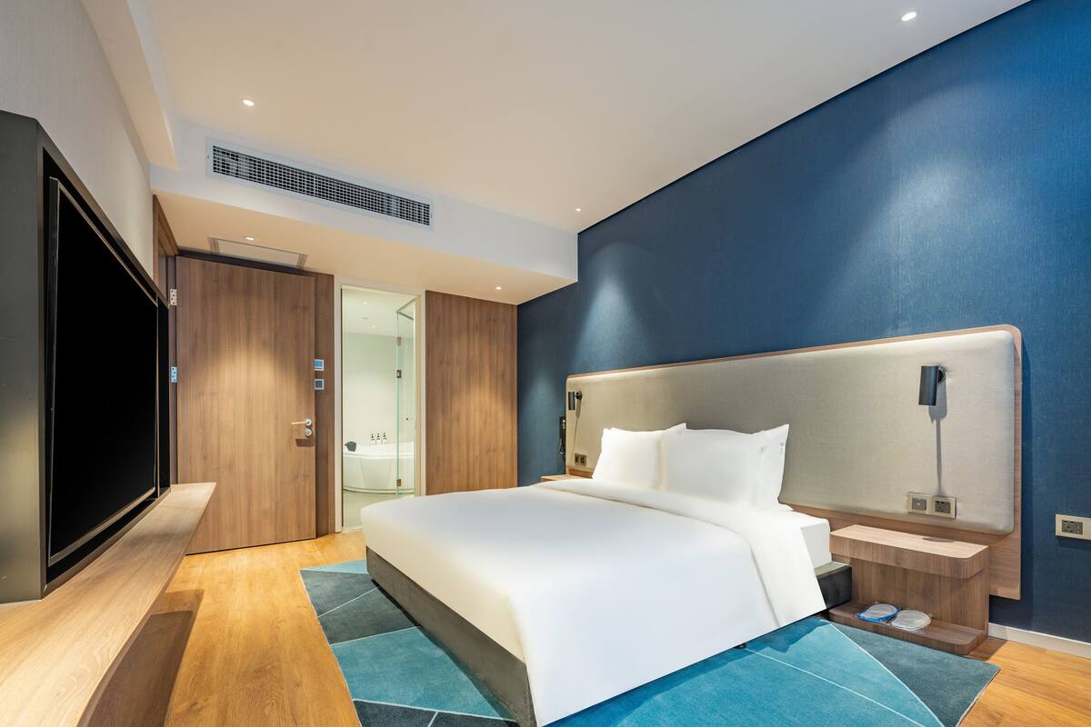 A modern hotel room with a large bed, a flat-screen TV, wooden flooring, and a blue accent wall.