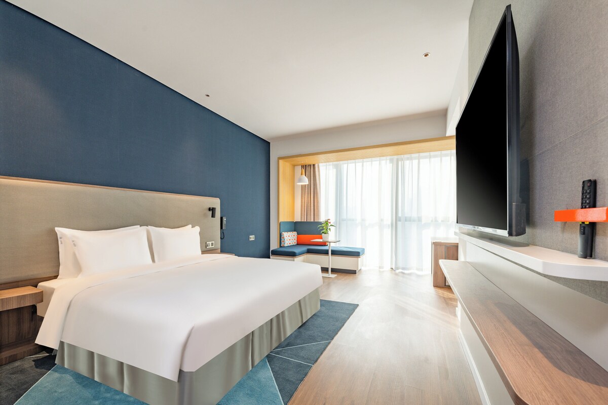 A modern hotel room with a large bed, a flat-screen TV mounted on the wall, a seating area with a table and chairs, and a large window with curtains.
