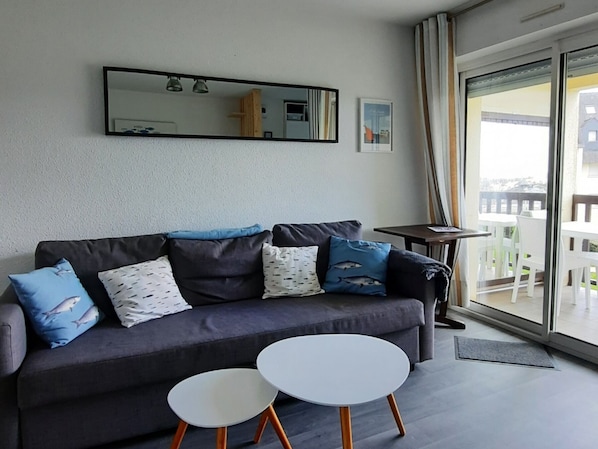 A modern living room with a grey sofa, two white round tables, and a large window with a view of a balcony.