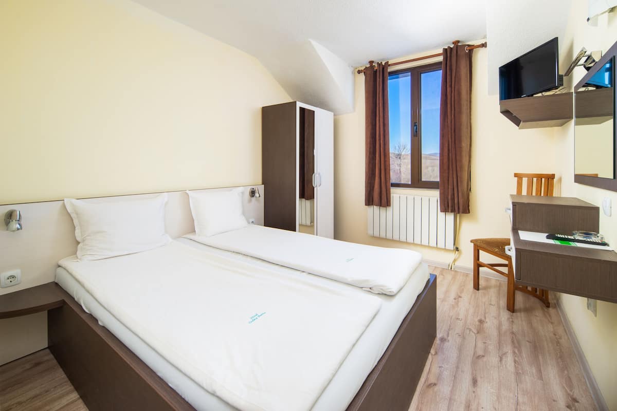 Een tweepersoonsbed met witte lakens, een houten bureau, een stoel, een televisie en een raam met gordijnen.