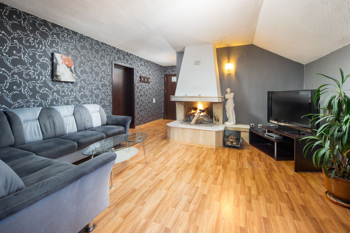 Een moderne woonkamer met een grijze bank, een glazen salontafel, een open haard, een televisie en een potplant.