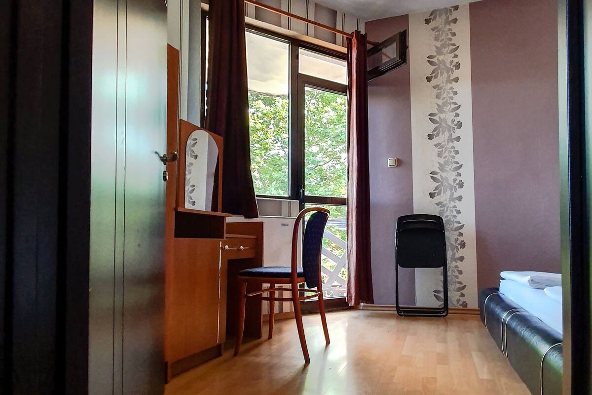 Een kamer met een houten bureau, een stoel, een bed, een raam met gordijnen en een muur met een bloemenmotief.