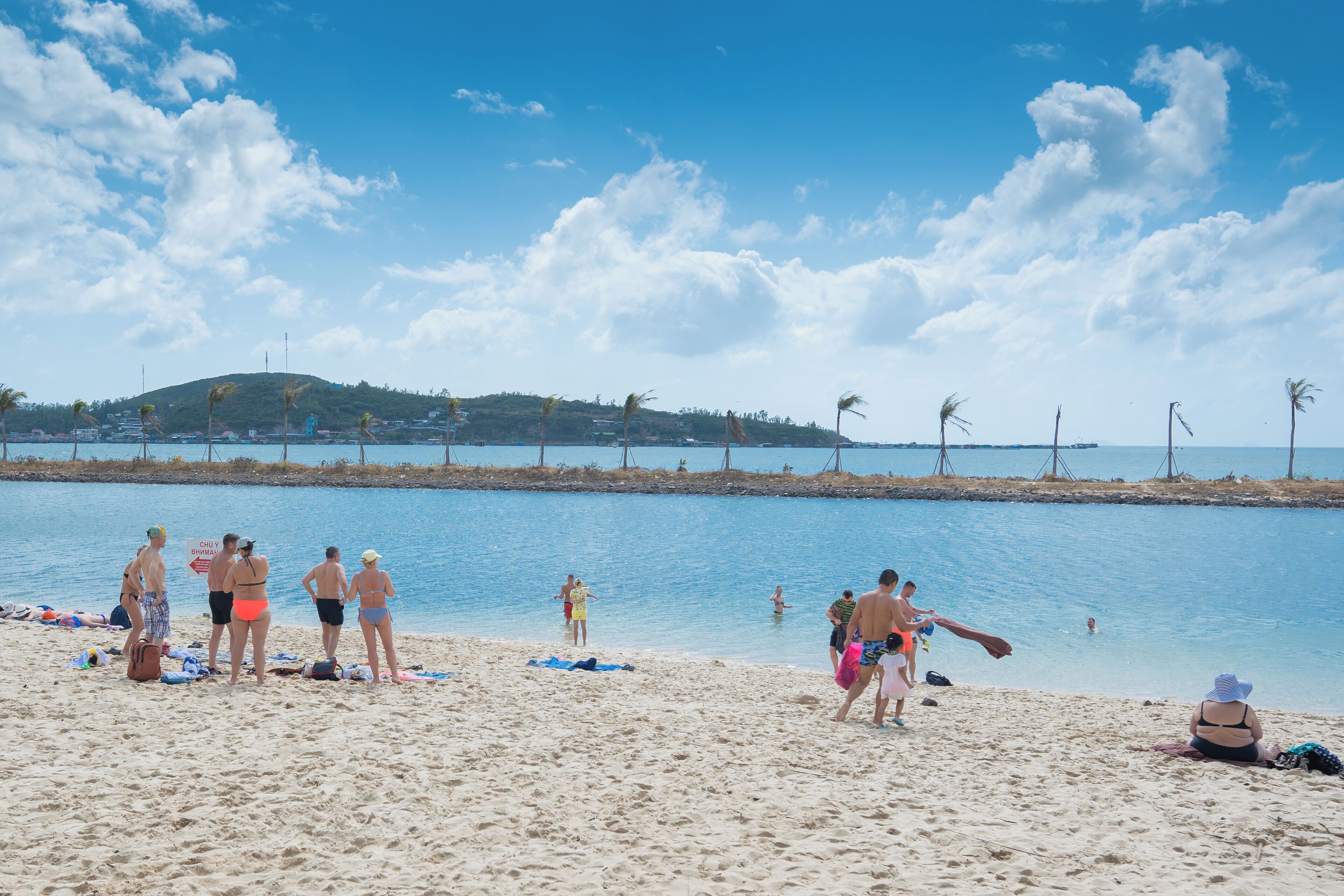 Uma praia com pessoas tomando sol e nadando, céu claro e uma colina distante com árvores.