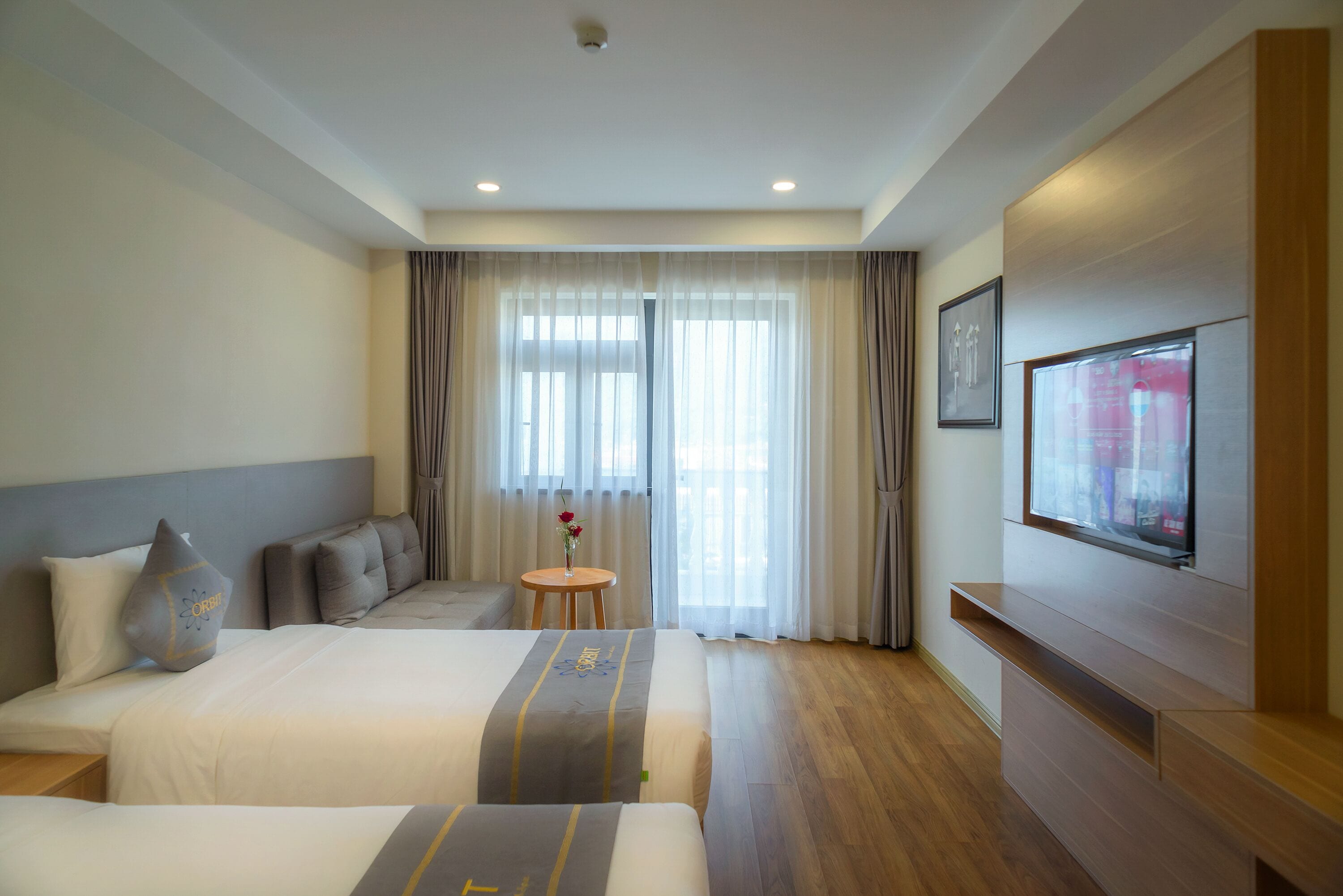 A hotel room with two beds, a small table with a vase, a TV on the wall, and a large window with curtains.