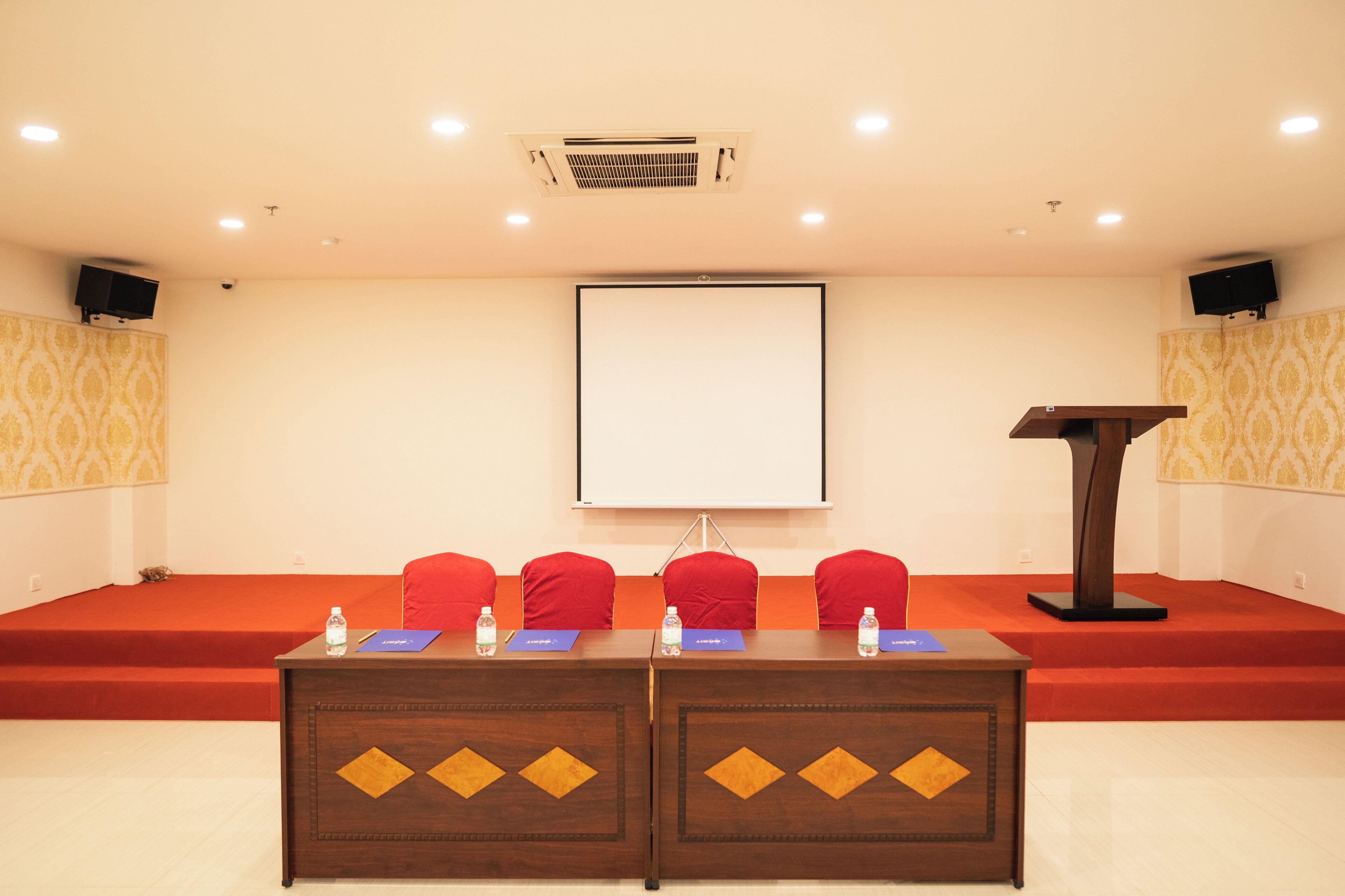 A conference room with a stage, red chairs, a podium, and a projector screen.