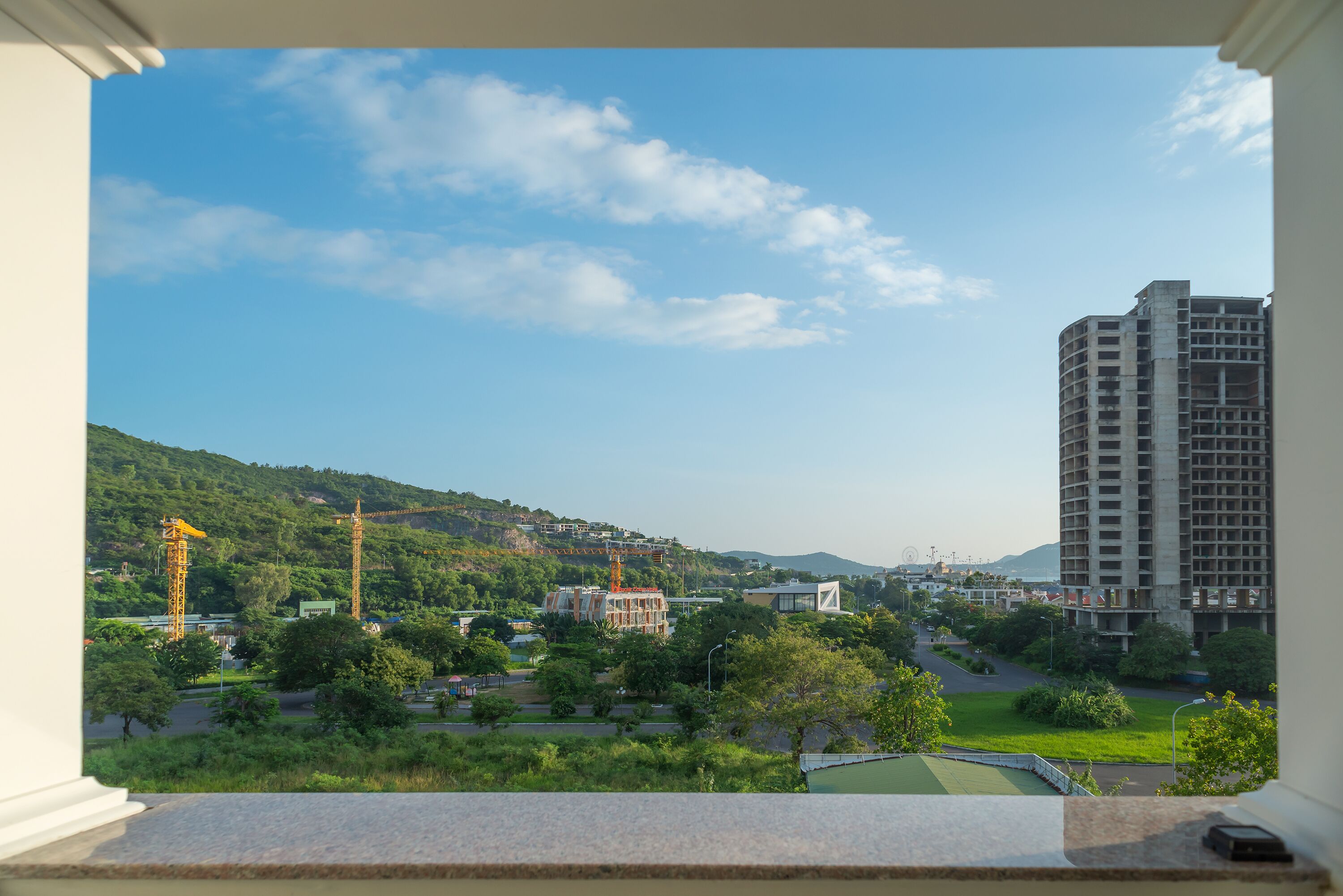 A view from a balcony overlooking a green landscape with trees, a road, and a building under construction.