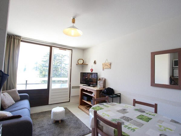 A room with a sofa, a small ottoman, a dining table with a green tablecloth, a TV, and a wooden desk.