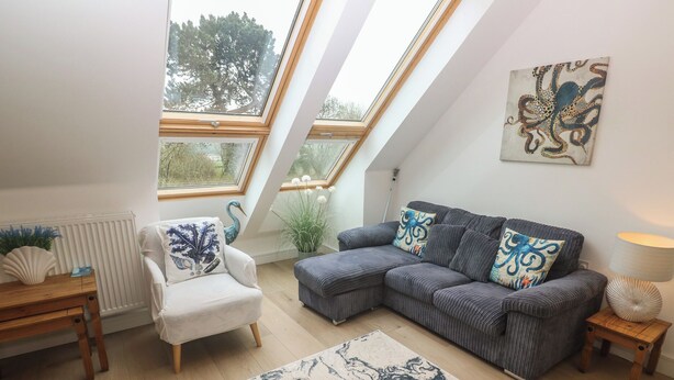 A loft-style room with a sloped ceiling, a grey sectional sofa, a white armchair, a wooden side table, and a large window with a view of trees. - Upper Deck (Truro)