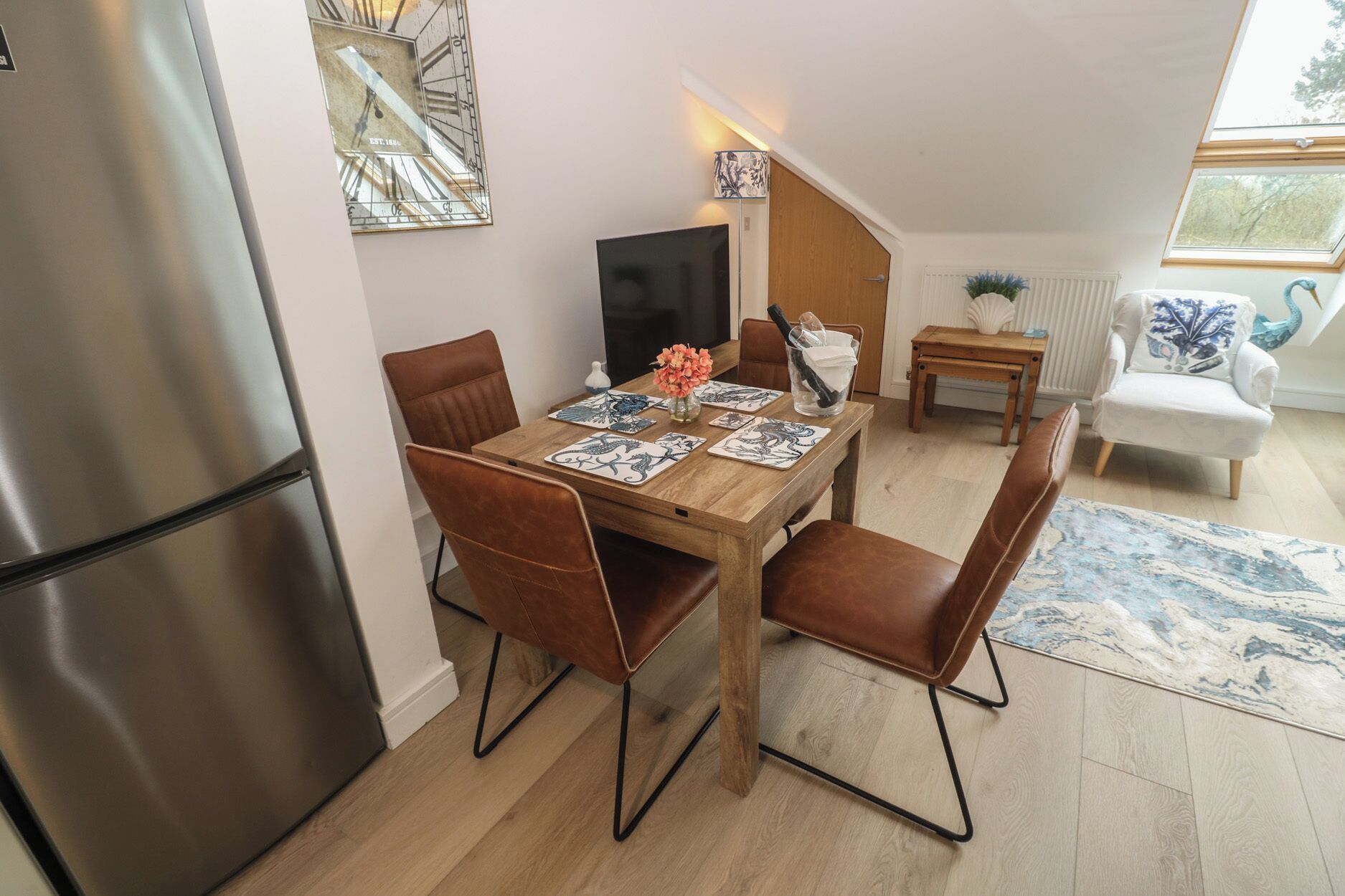 Uma área de refeições com mesa de madeira, cadeiras marrons, uma televisão e uma pequena mesa lateral com um vaso e uma luminária.