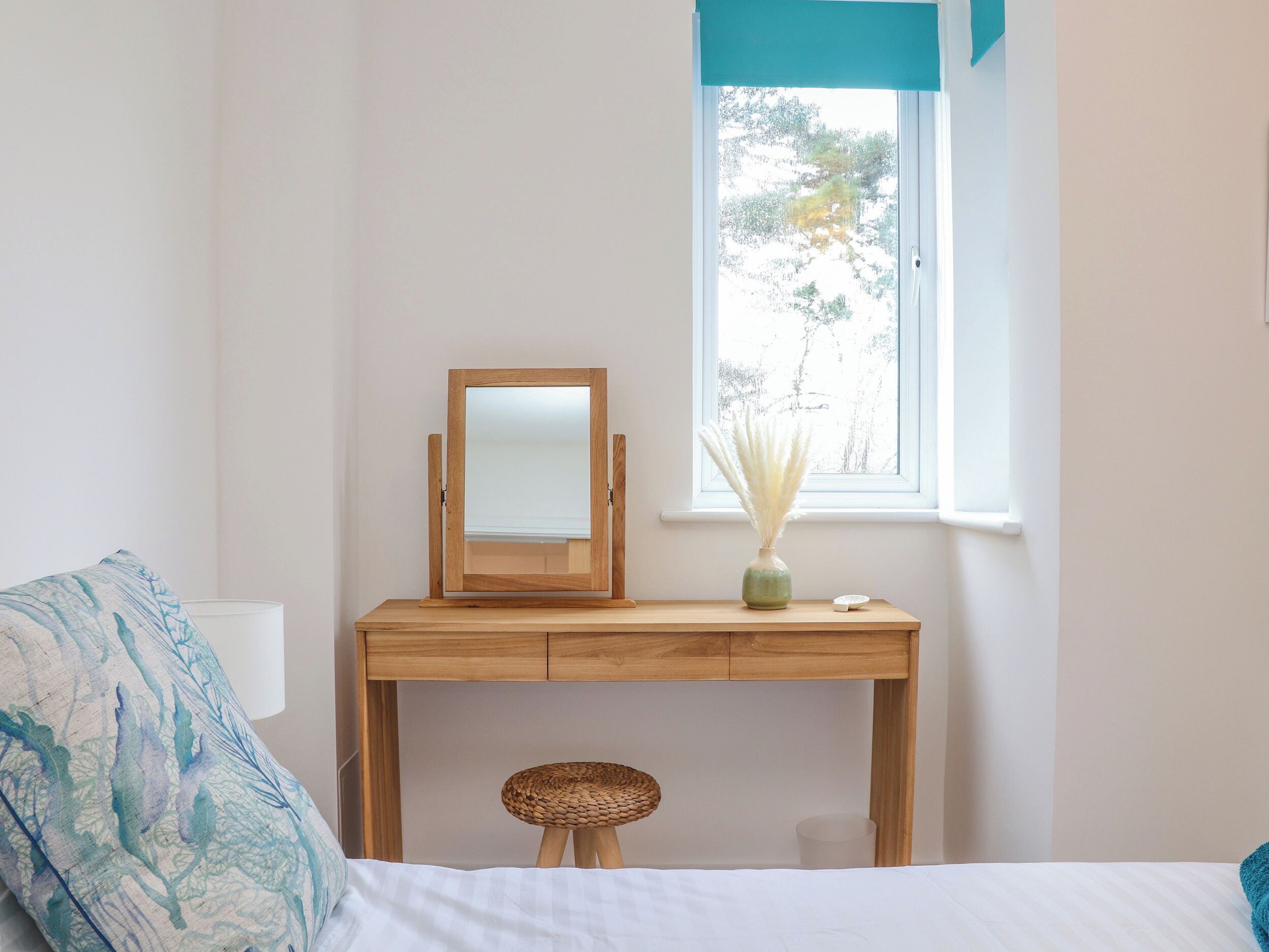 Un dormitorio con una cama, un escritorio de madera con un espejo, un taburete de mimbre y una ventana con vistas a árboles.
