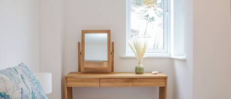 Un dormitorio con una cama, un escritorio de madera con un espejo, un taburete de mimbre y una ventana con vistas a árboles.