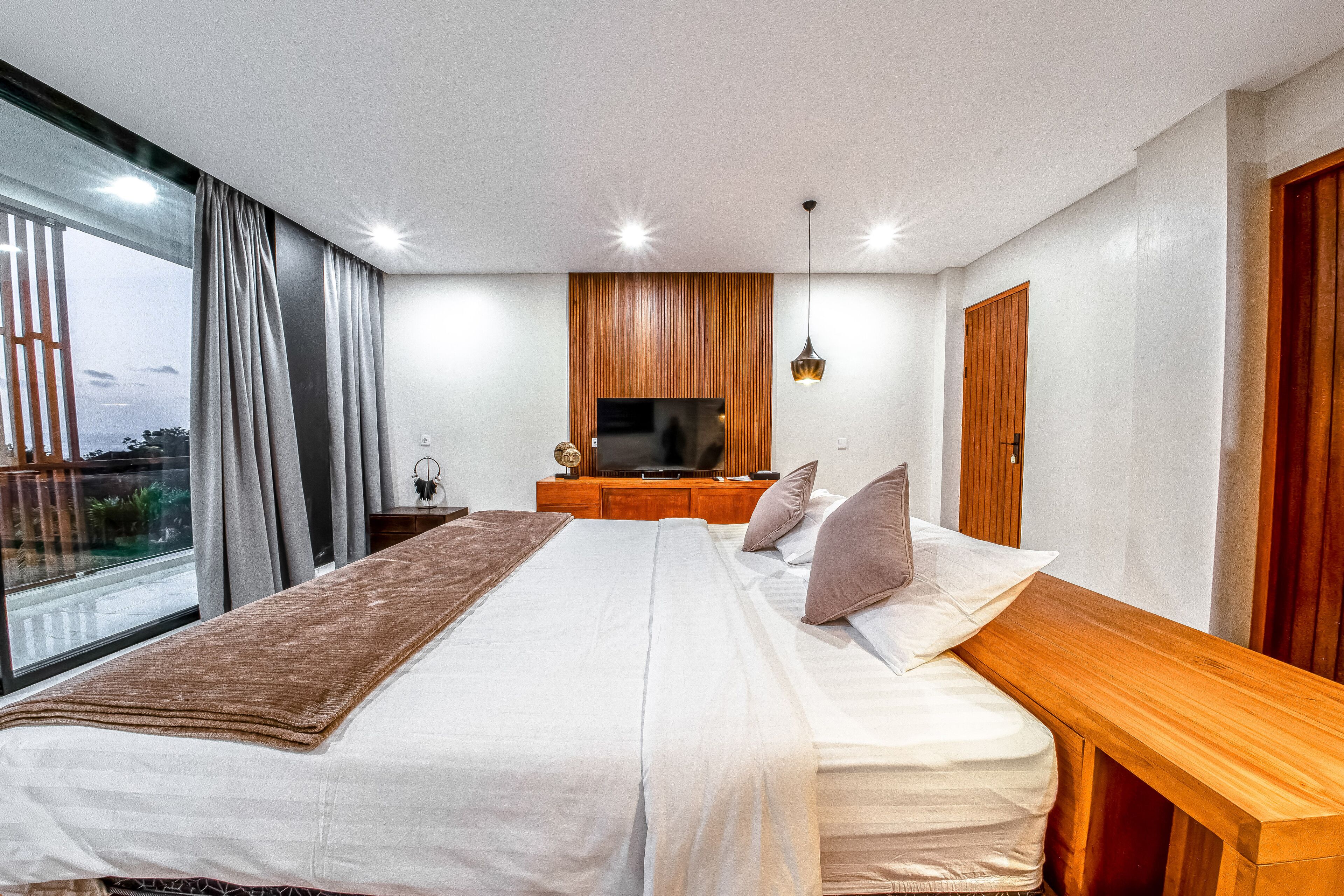 Une chambre moderne avec un grand lit, une tête de lit en bois, une télévision et une vue sur l’extérieur.