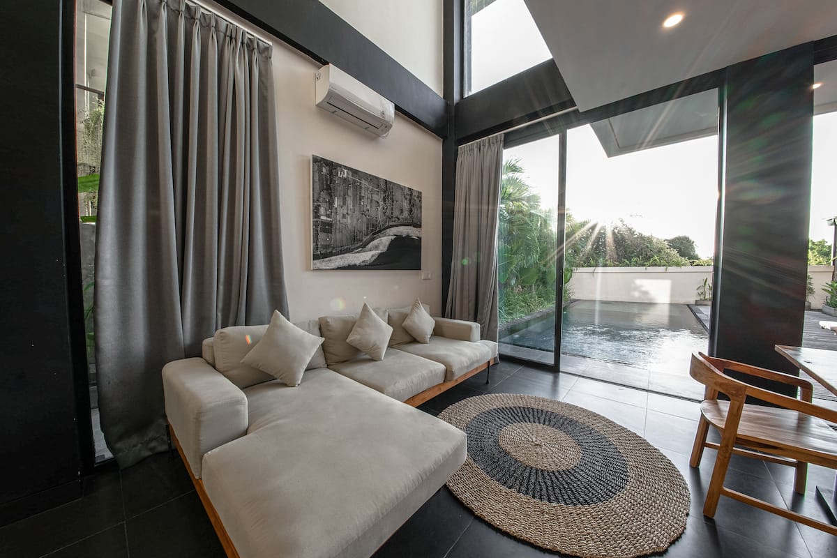 Ein modernes Wohnzimmer mit einem Ecksofa, einem Esstisch mit Stühlen und einem großen Fenster mit Vorhängen.