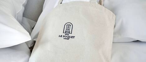 Un sac fourre-tout en toile de couleur beige, orné du logo et du nom d’un hôtel, posé sur un lit recouvert de draps blancs.