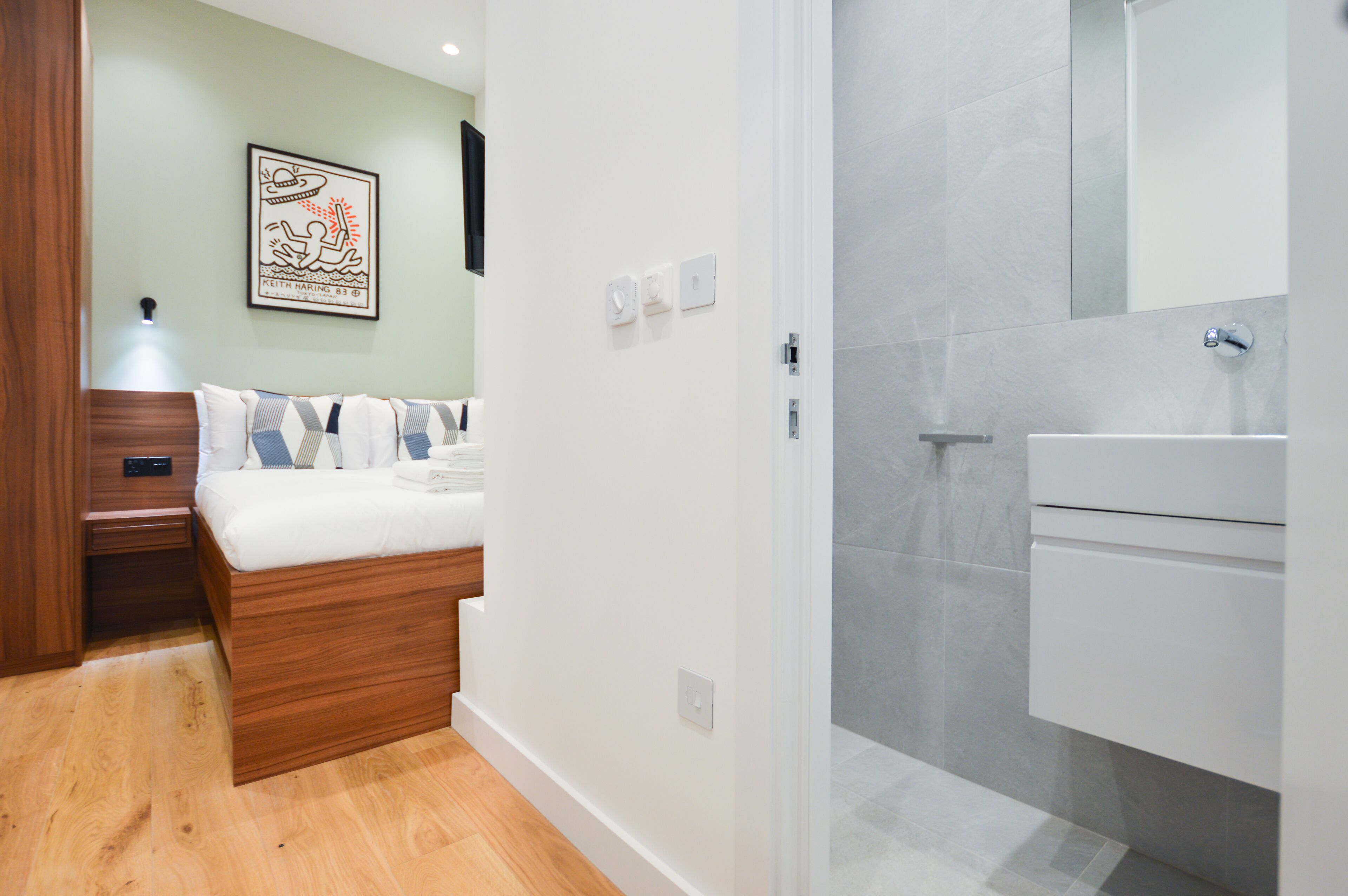Une chambre moderne avec un lit en bois, une table de chevet et une commode. La salle de bain est équipée d’un lavabo blanc et d’un espace douche.