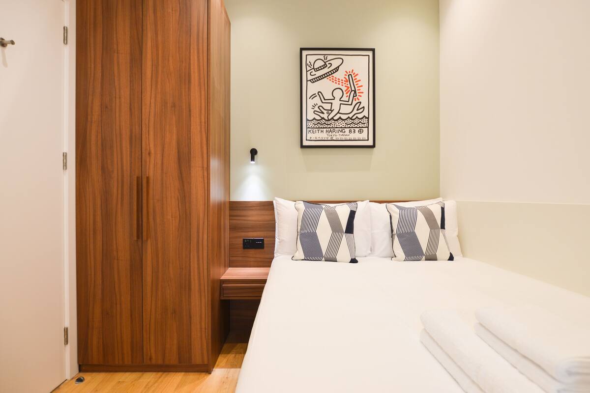 A bedroom with a wooden wardrobe, a bed with patterned pillows, and a framed artwork on the wall.