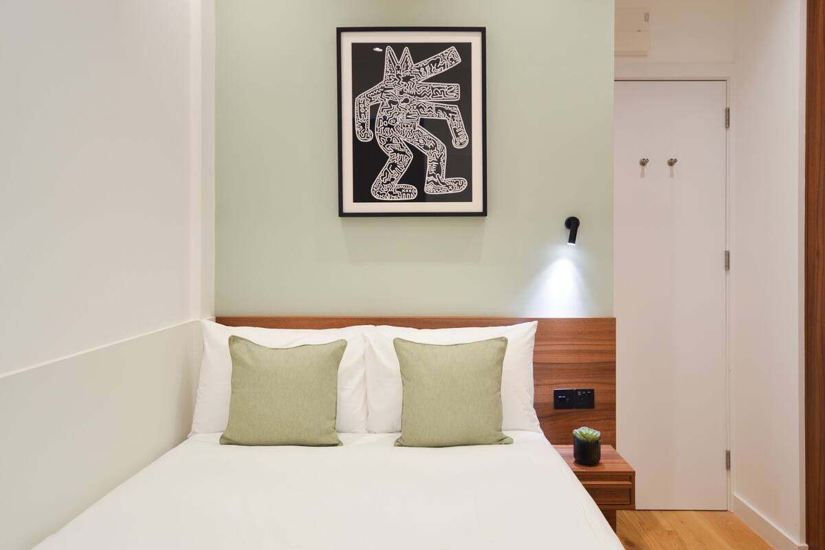 A neatly made bed with two pillows, a framed artwork on the wall, and a bedside table with a small plant.