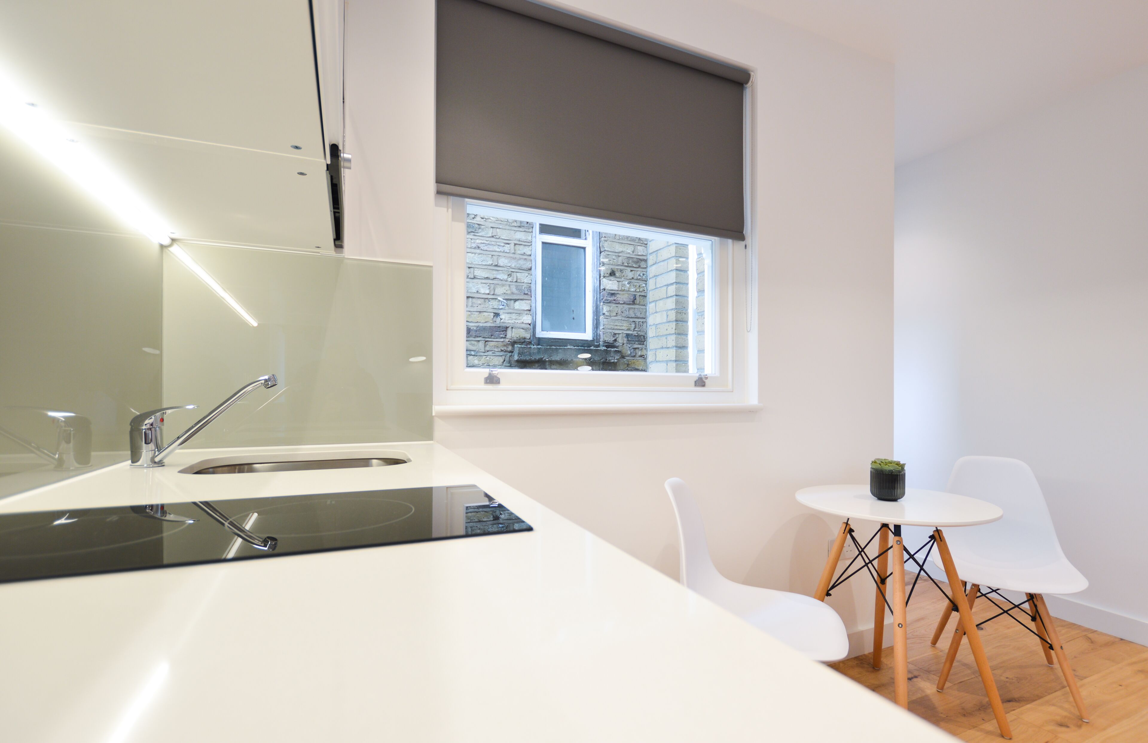 Cocina moderna con estufa integrada, lavamanos y ventana con vista a un edificio de piedra. Hay una pequeña mesa con sillas y una planta en maceta.