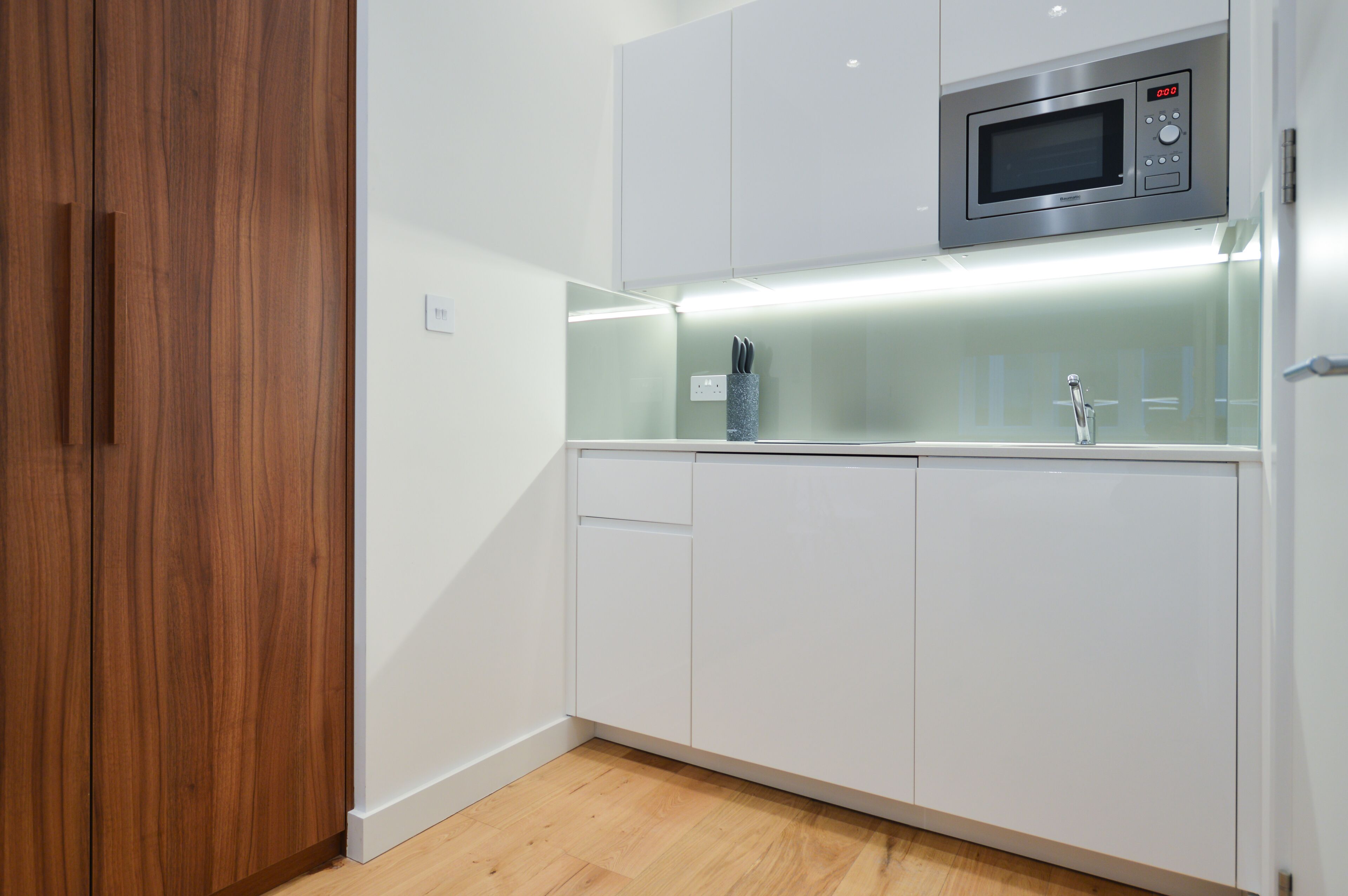 Een moderne keuken met witte kastjes, een magnetron en een glazen spatwand.