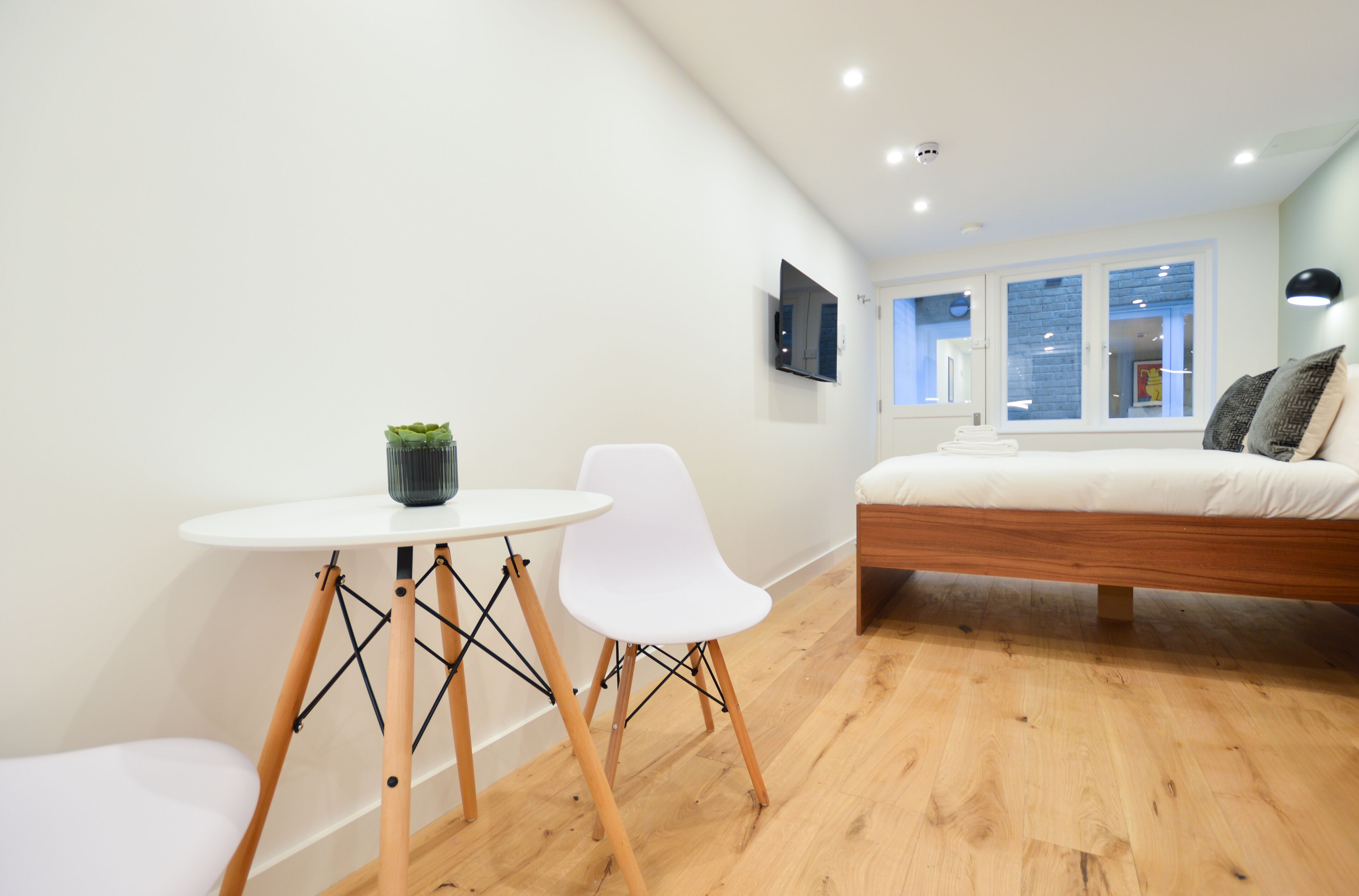 Une chambre moderne avec un lit, une petite table sur laquelle est posée une plante, une chaise blanche et une fenêtre donnant sur la ville.