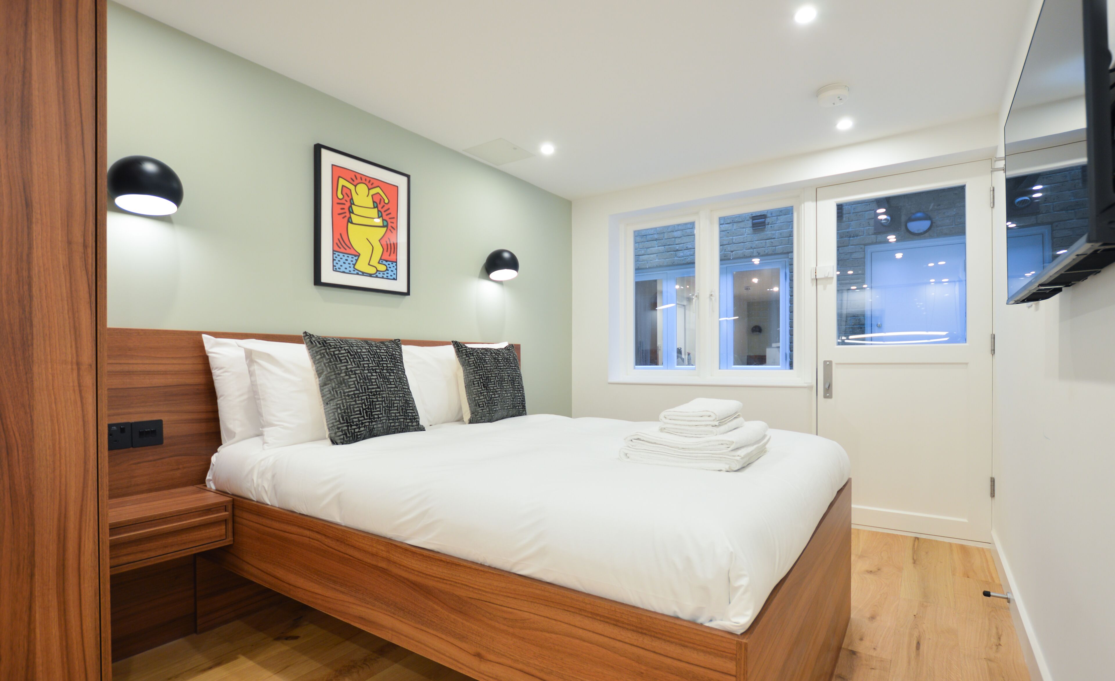 A modern bedroom with a wooden bed, a framed artwork, two wall-mounted lights, and a window with a view of the interior.