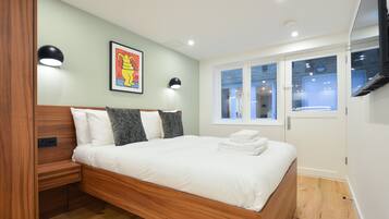 A modern bedroom with a wooden bed, a framed artwork, two wall-mounted lights, and a window with a view of the interior.