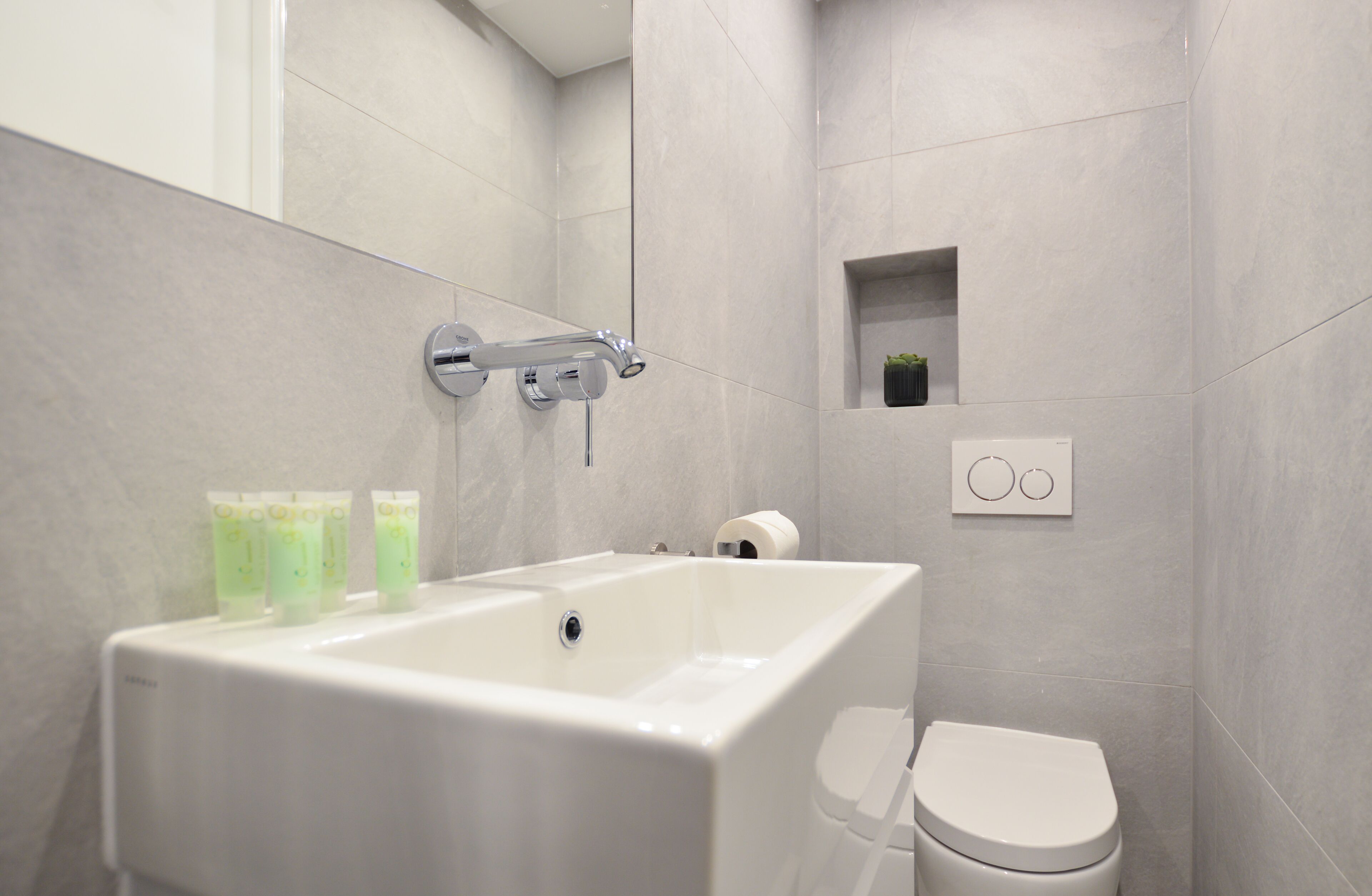 A modern bathroom with a white bathtub, toilet, and a small shelf with toiletries.