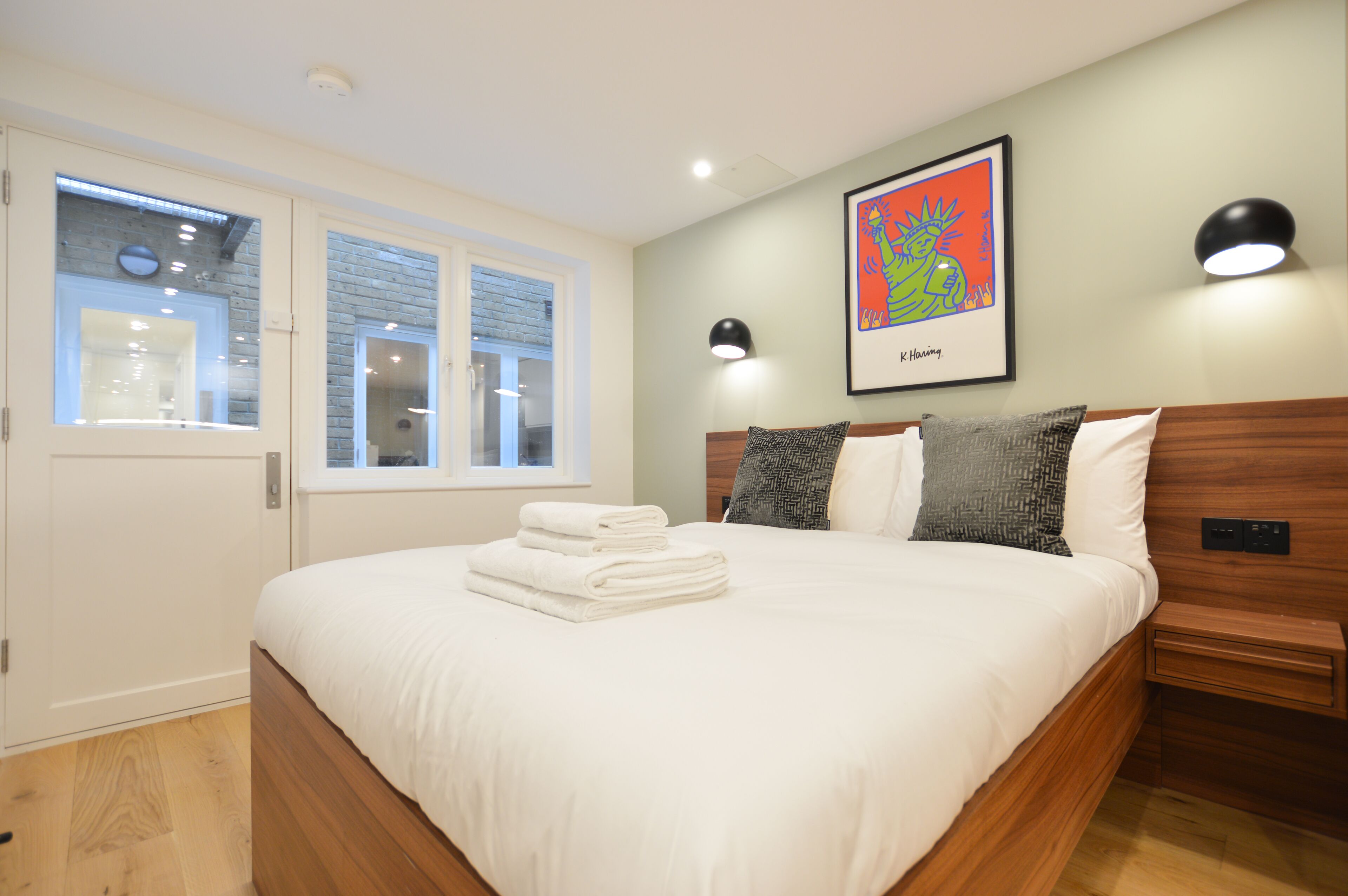 A neatly made bed with white linens and a folded towel, a wooden headboard, a wall-mounted artwork, and a bedside table with a lamp.