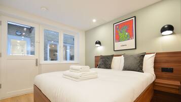 A neatly made bed with white linens and a folded towel, a wooden headboard, a wall-mounted artwork, and a bedside table with a lamp.