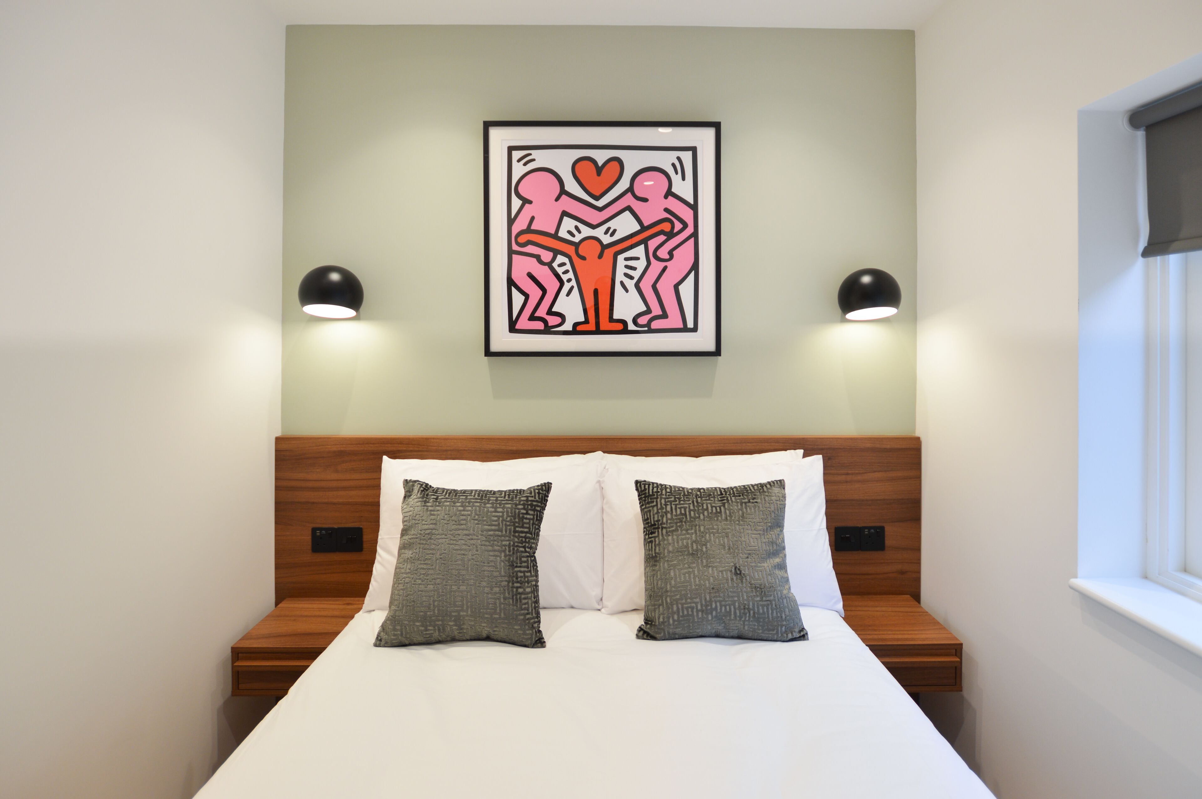Een netjes opgemaakt bed met twee kussens, een lijstwerk erboven en bedlampjes.