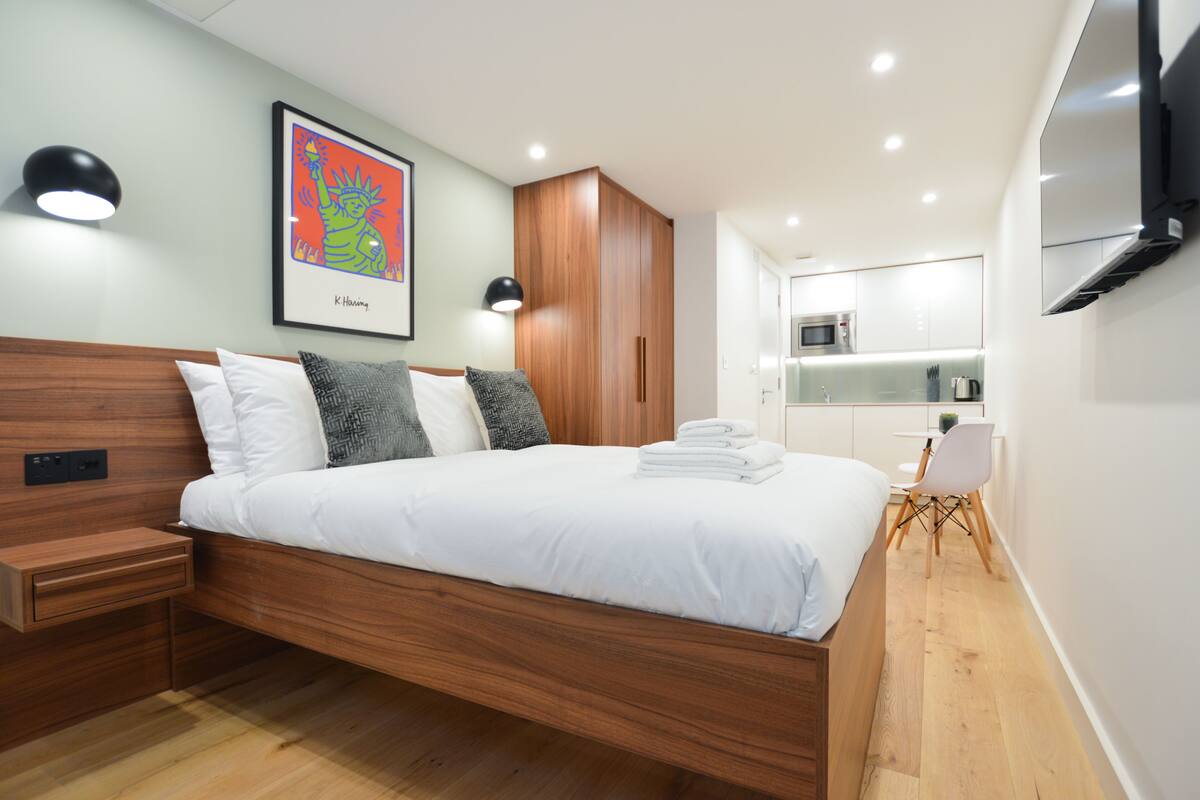 A modern bedroom with a wooden bed, bedside tables, a wall-mounted TV, and a kitchen area in the background.