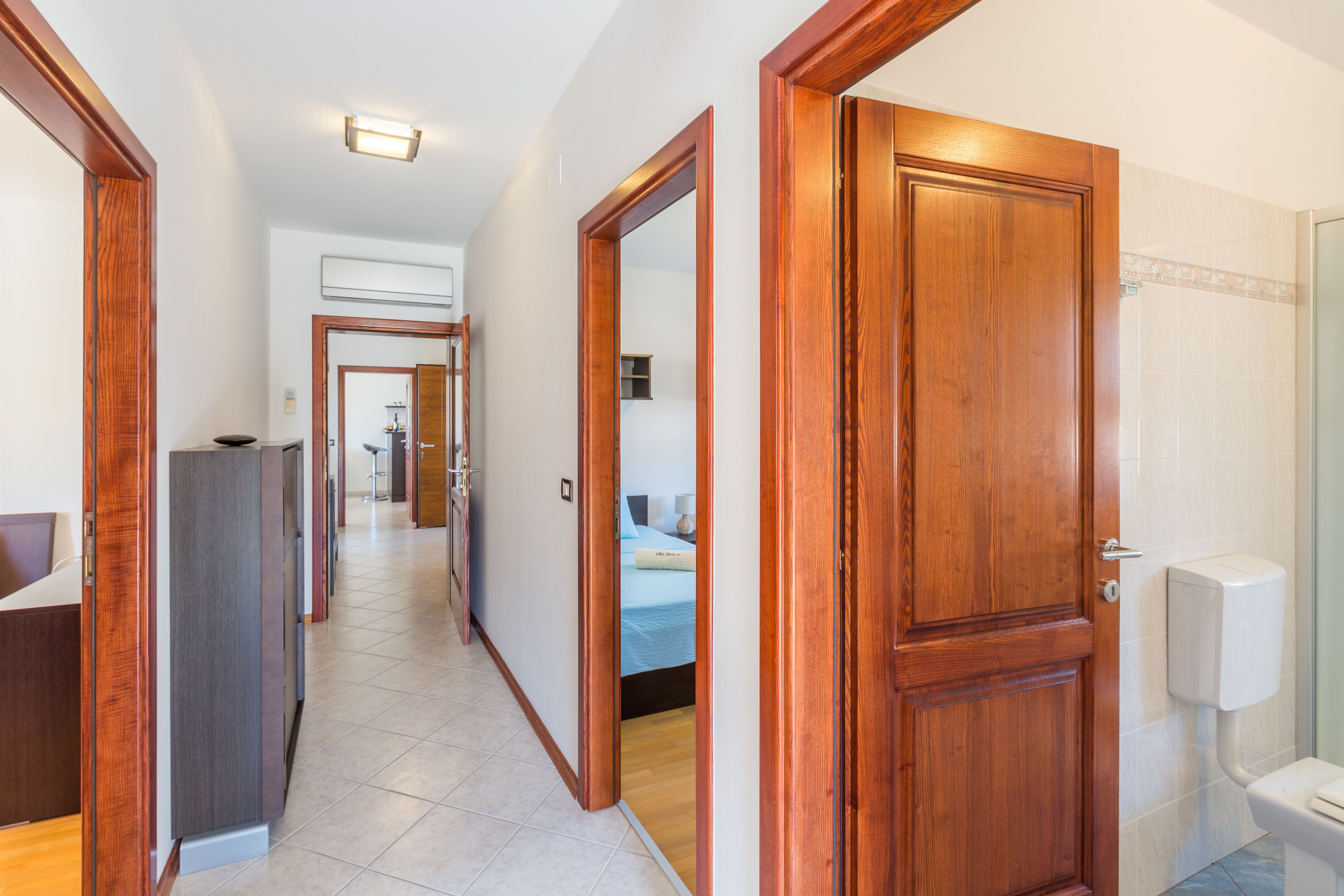 A hotel hallway with wooden doors, a bed in the background, and a bathroom with a toilet and shower.