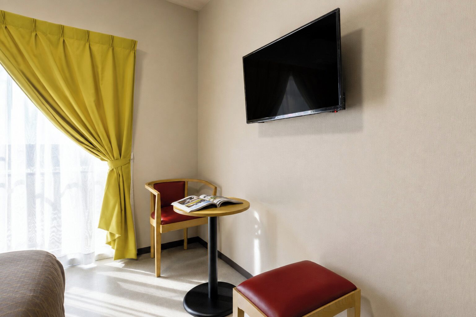 Quarto de hotel com televisão na parede, uma mesa redonda pequena com uma revista, uma cadeira vermelha e uma cortina amarela.