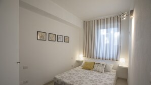 A bedroom with a bed, a window with curtains, a wall with framed pictures, and a nightstand with a lamp. - BON Repos Apartment With Swimming Pool (Alghero)