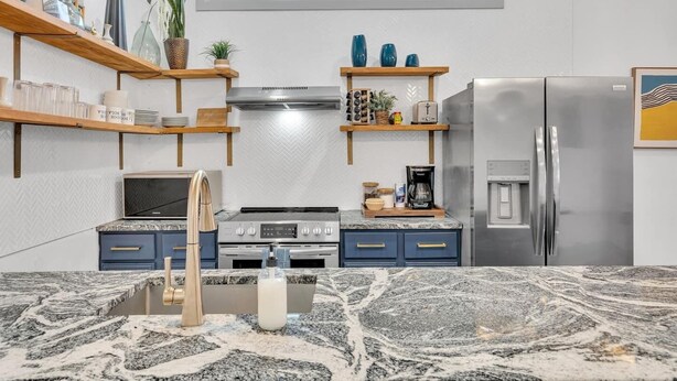 A modern kitchen with a gas stove, oven, and stainless steel refrigerator. There are wooden shelves with decorative items and a window above the sink.