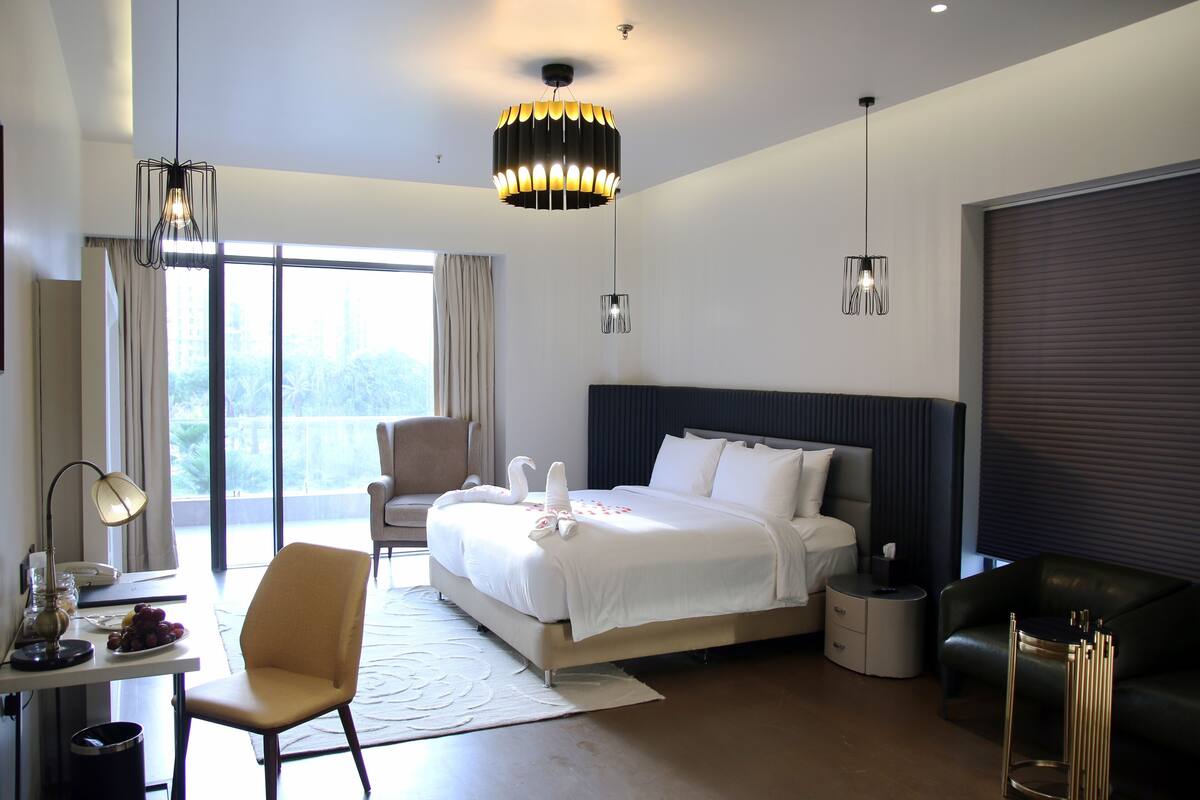 Ein modernes Hotelzimmer mit einem großen Bett, einem Schreibtisch mit Lampe, einem Sessel, einer Couch und Blick ins Freie.