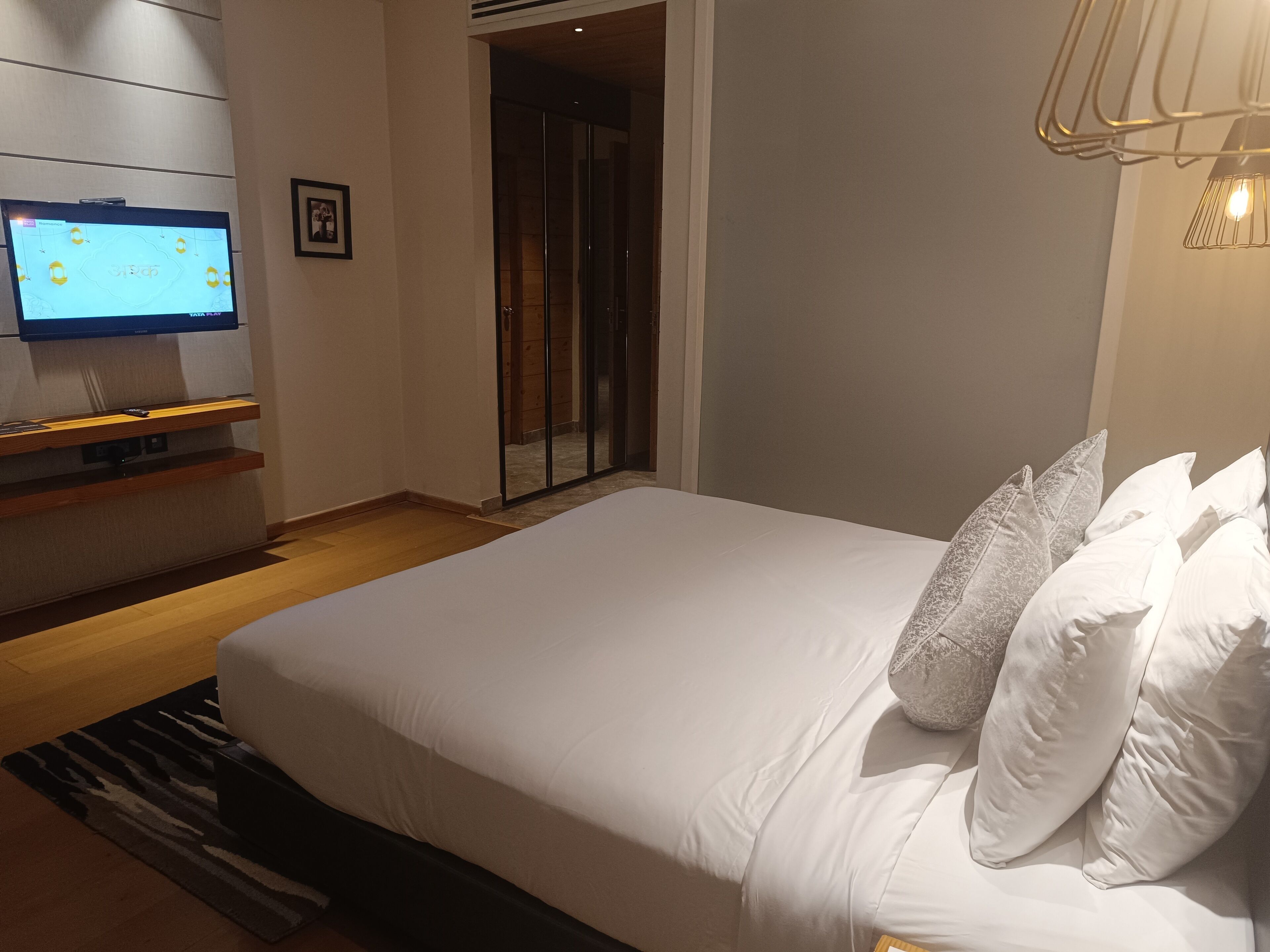 Una habitación de hotel moderna con una cama grande, un televisor de pantalla plana montado en la pared, piso de madera y una puerta de vidrio corredera que da a un balcón.