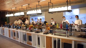 Une cuisine moderne où des chefs préparent des plats, avec un plafond en lambris de bois et des luminaires contemporains.