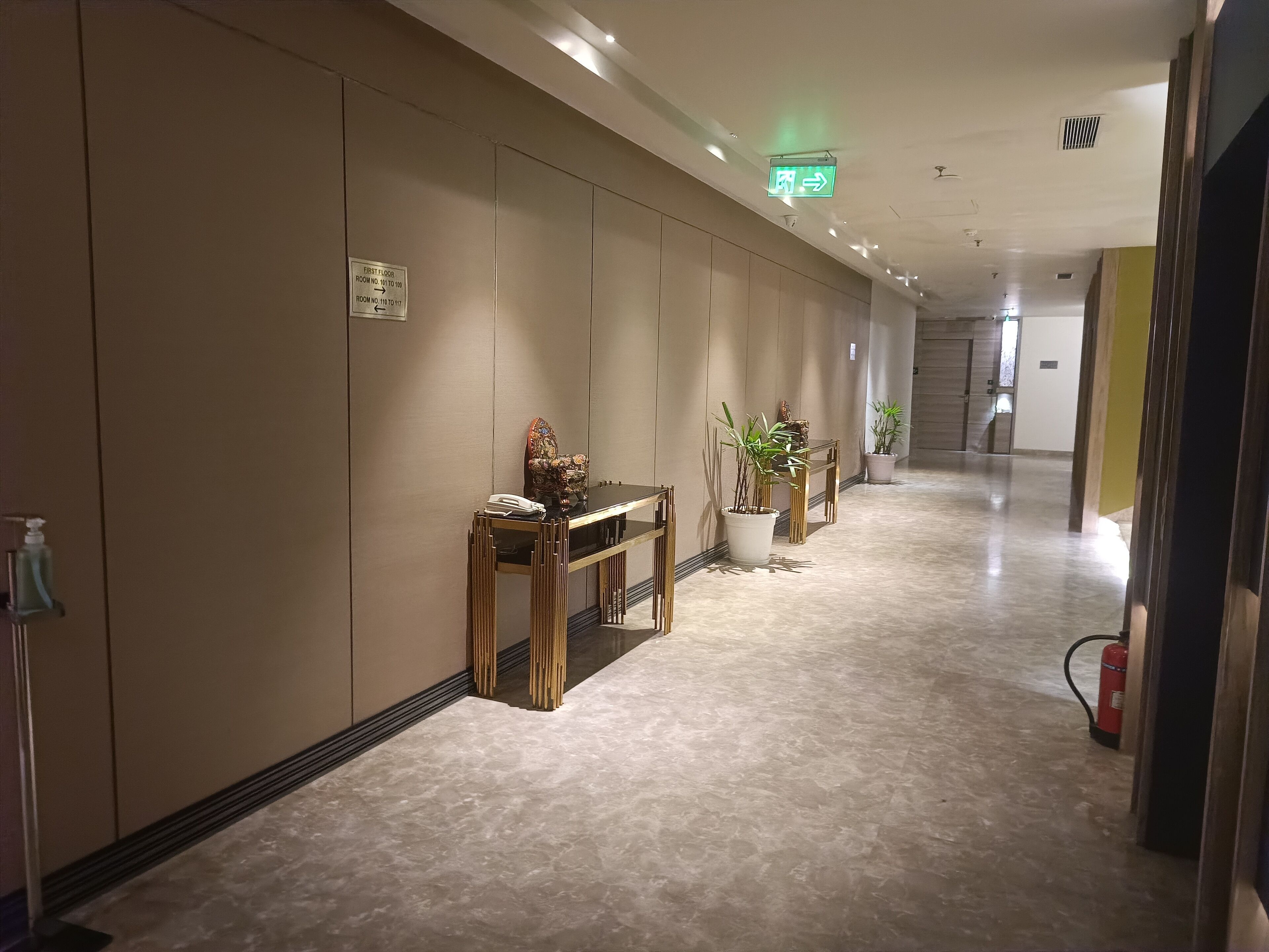 A hotel corridor with a marble floor, a reception desk with a telephone, a decorative chair, and potted plants.