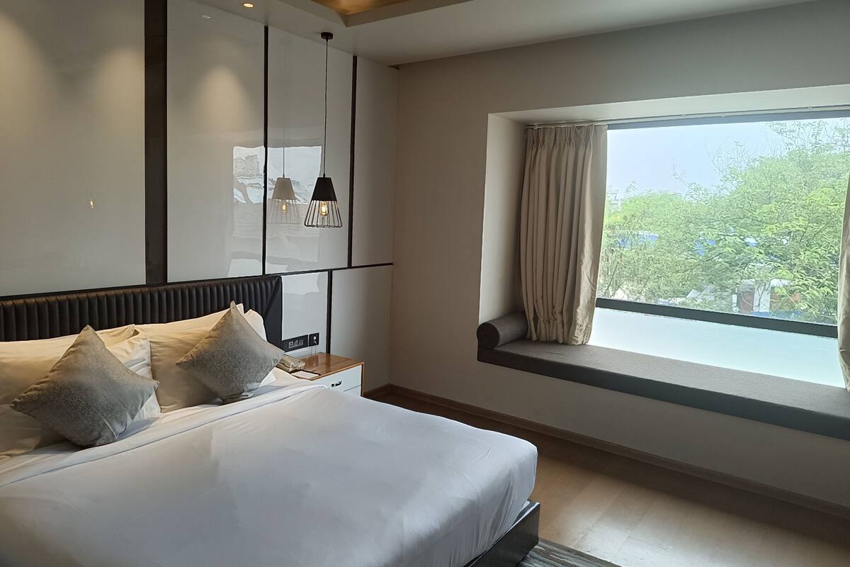 Ein modernes Schlafzimmer mit einem großen Bett, einem Nachttisch, einem Fenster mit Vorhängen und Blick ins Grüne.