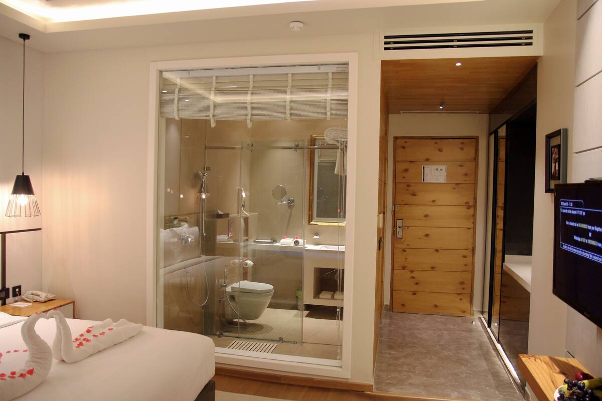 Ein Hotelzimmer mit einer gläsernen Dusche, einem Bett mit weißem Handtuch, einer Holztür und einem an der Wand befestigten Fernseher.