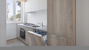 A modern kitchen with a built-in dishwasher, stainless steel appliances, and a window with a view of palm trees. - Italianway - Cristoforo Colombo 12 INT 1A (San Bartolomeo al Mare)