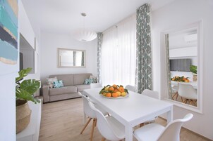 Un comedor moderno y luminoso con una mesa y sillas blancas, un sofá con cojines estampados, y un espejo grande que refleja la habitación.
