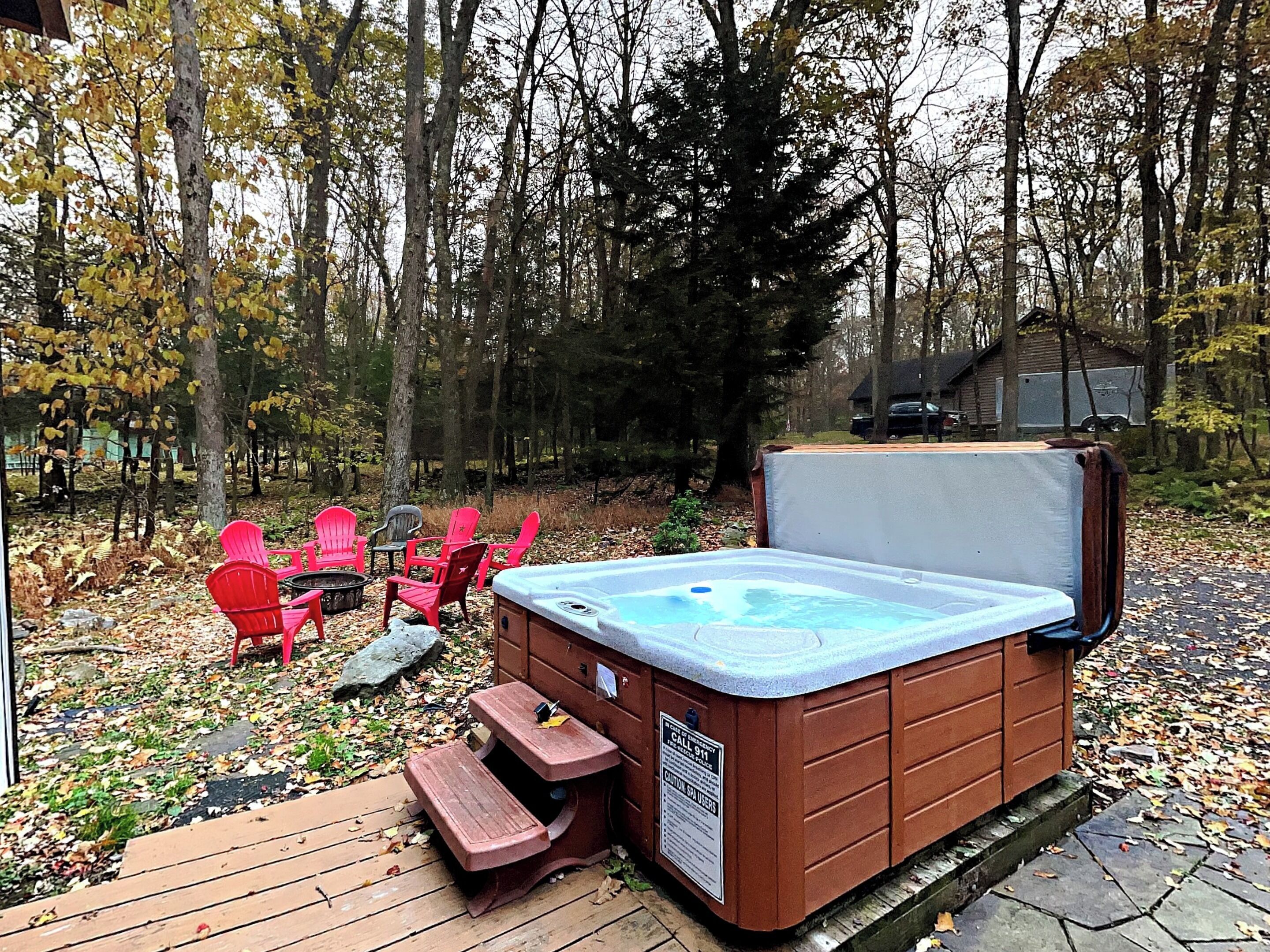 A hot tub with a wooden deck, surrounded by trees and outdoor seating.