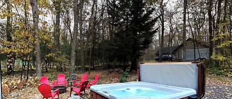 A hot tub with a wooden deck, surrounded by trees and outdoor seating.
